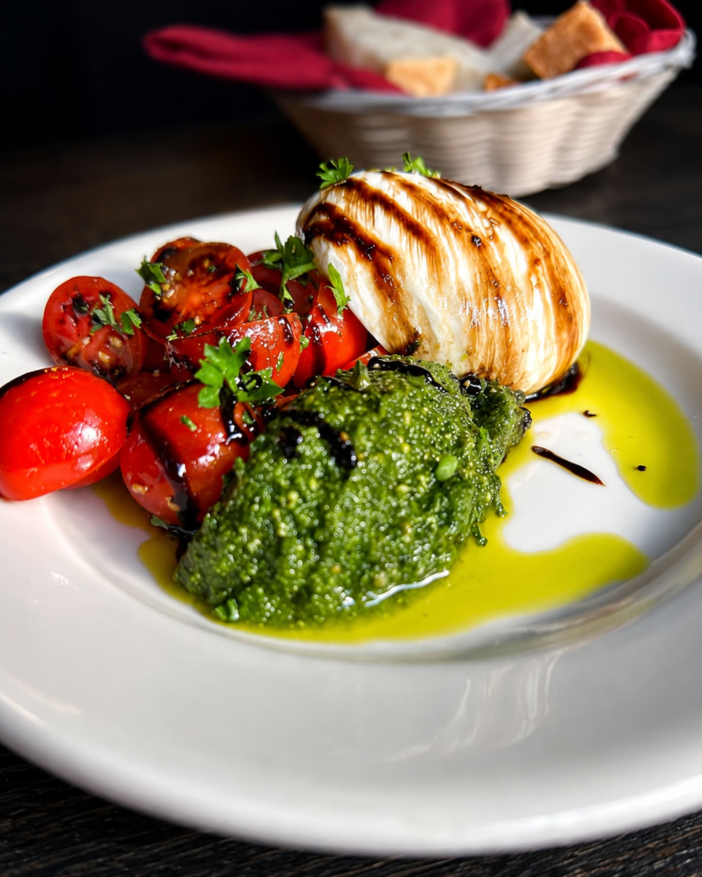 A ball of burrata on a white plate beside blistered cherry tomatoes and a spoonful of bright green basil pesto, with balsamic and olive oil pooled around them.