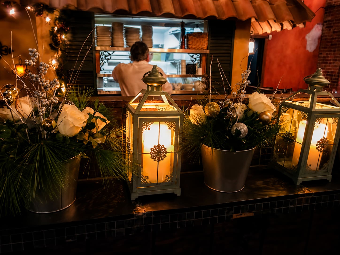 Candle lanterns and planted flowers along a ledge in the courtyard, with the kitchen lit up through a window behind them and a cook working inside.