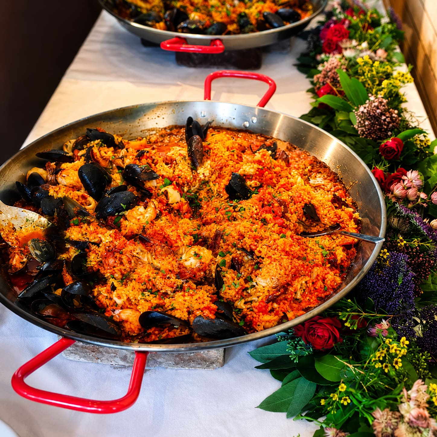 A steel paella pan with red handles, full of saffron rice with black mussel shells through it, resting on a table edged with flowers.
