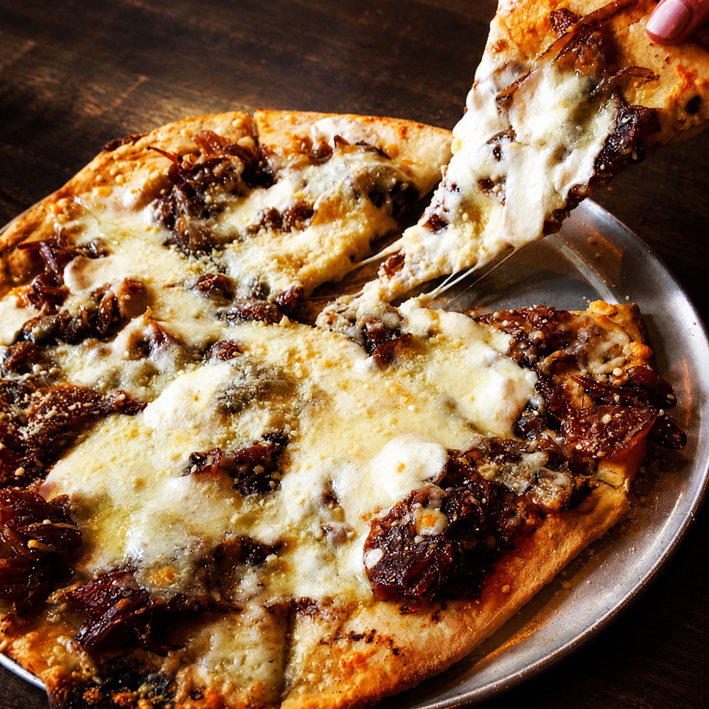 A thin-crust pizza on a metal pan, blistered at the edges, with a slice being lifted and the melted cheese pulling away in strands.