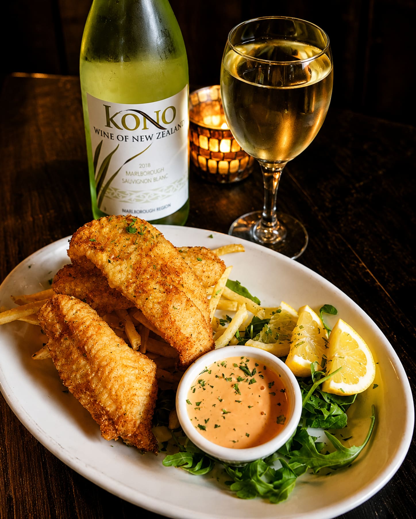 Crisp fried fish fillets with hand-cut fries, lemon wedges and a cup of citrus aioli, beside a poured glass of white wine.
