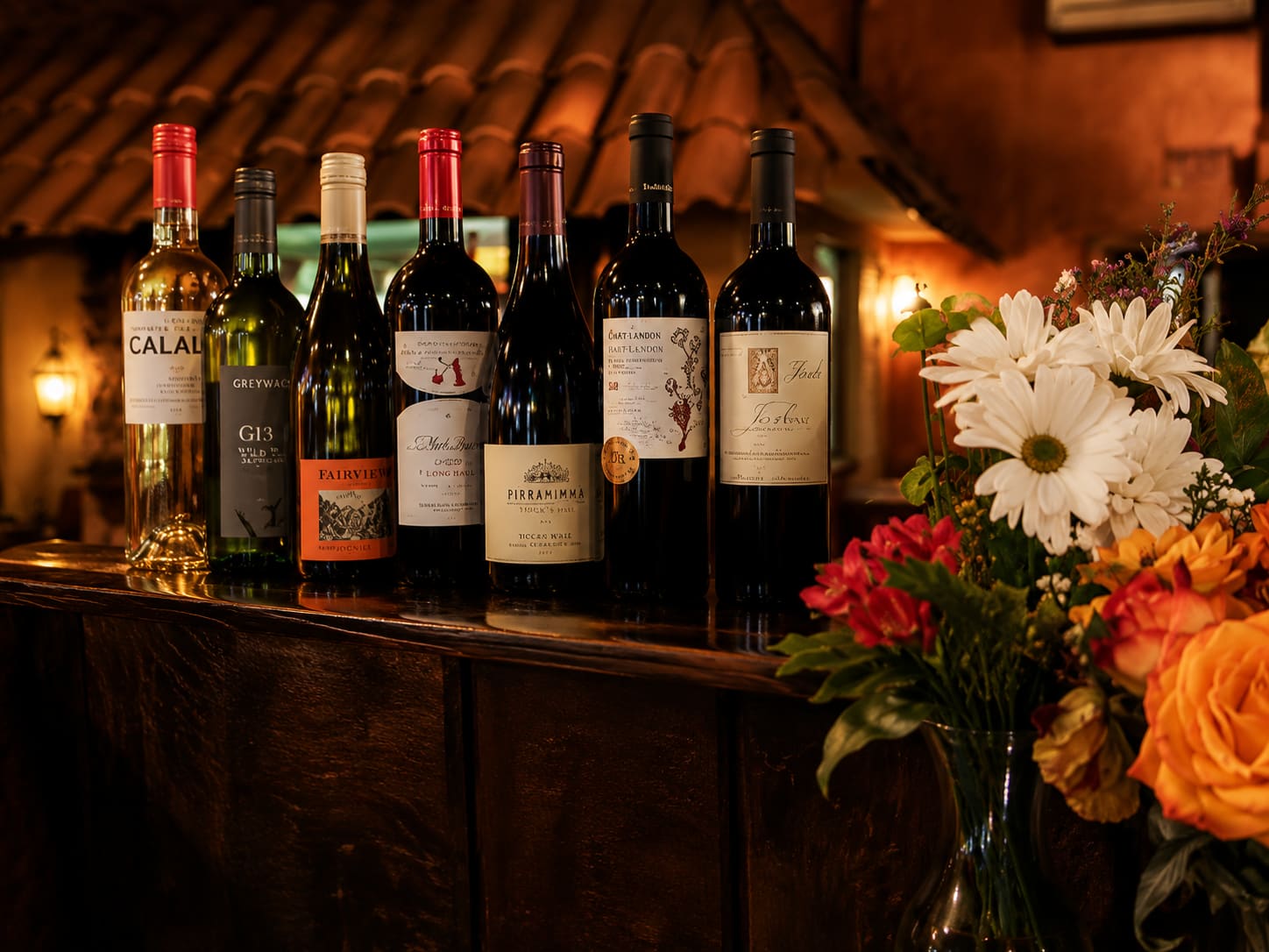 A row of wine bottles standing along a wooden ledge in warm low light, with cut flowers in front of them.